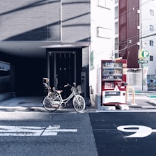 A bicycle with a child's seat is parked next to a vending machine on a city street. The background consists of urban buildings and some parking signage.