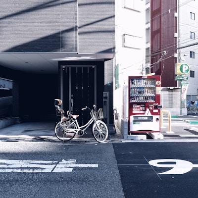 A bicycle with a child's seat is parked next to a vending machine on a city street. The background consists of urban buildings and some parking signage.
