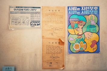 A collection of vintage Korean posters attached to a wall. The poster on the left features cartoon illustrations and text related to ramen flavors from different years. In the center, there are two aged, paper-like documents containing Korean text, with one having a visible crease. On the right, a colorful illustration shows a girl, a tree with flowers, a hand reaching for a bag, and a bird, accompanied by Korean text.