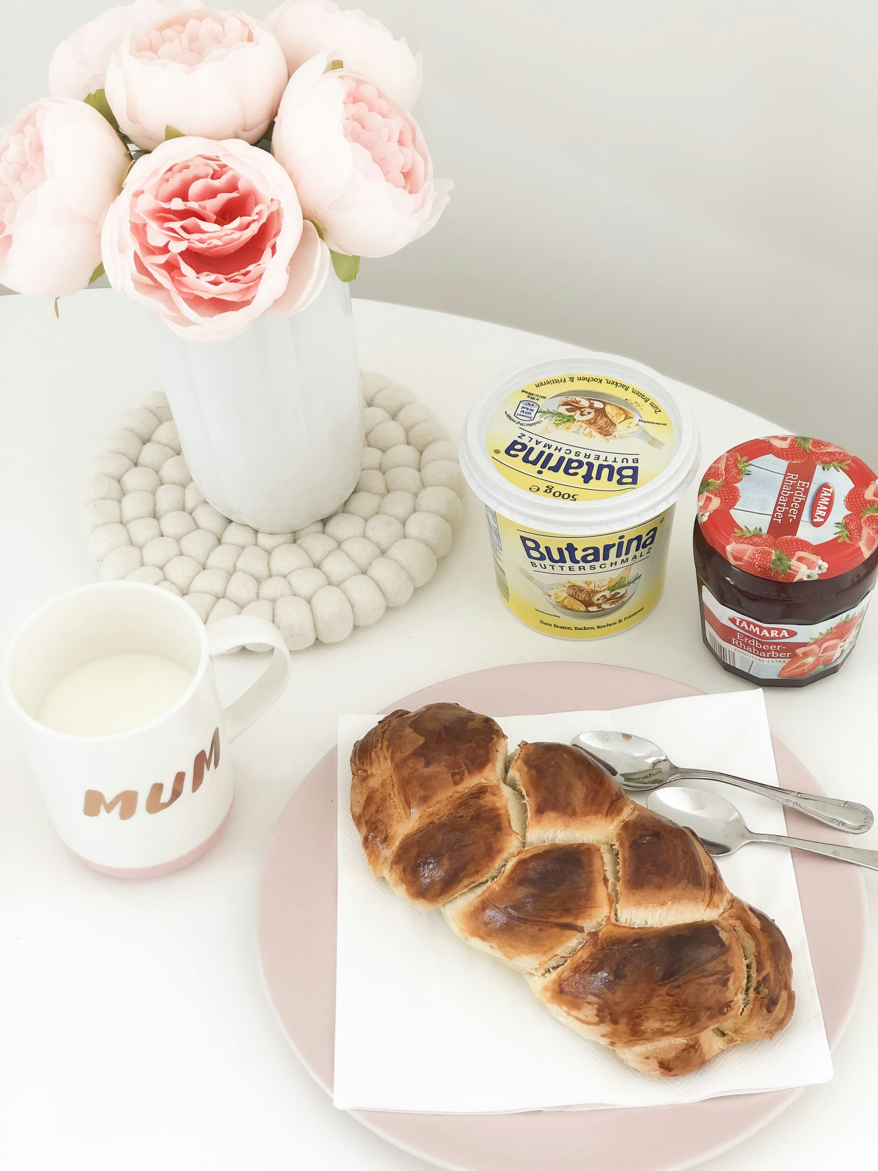 baked bread near spoon on plate beside milk in white mug and pink rose flowers in vase