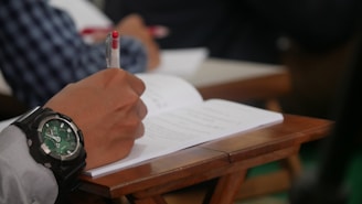 A person is writing in a notebook with a red pen while seated at a wooden desk. The focus is on their hand, which is adorned with a large wristwatch featuring a green face. The background includes another person wearing a checkered shirt.