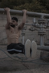 A shirtless man with a beard is performing a hanging exercise by gripping onto a large metal pipe. He is wearing a cap, black pants with a light-colored stripe, and sneakers. The background features industrial equipment and a concrete wall with foliage visible at the top.