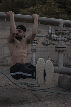 A shirtless man with a beard is performing a hanging exercise by gripping onto a large metal pipe. He is wearing a cap, black pants with a light-colored stripe, and sneakers. The background features industrial equipment and a concrete wall with foliage visible at the top.