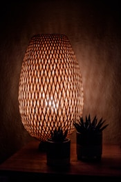 A wicker lamp emits a warm glow, creating intricate geometric patterns with its lattice design. The lamp is placed on a wooden surface surrounded by two small potted succulents.