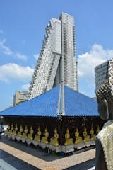 A modern high-rise building with a distinct architectural design featuring geometric patterns stands prominently against a clear blue sky. In the foreground, an array of golden Buddha statues are lined up along the side of a structure with a blue tiled roof. Part of a decorated figure is visible on the right, possibly another Buddha or a similar deity.