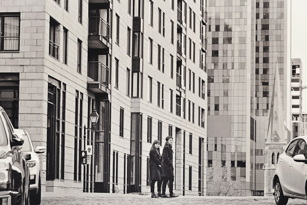 grayscale photo of man and woman standing on sidewalk near building