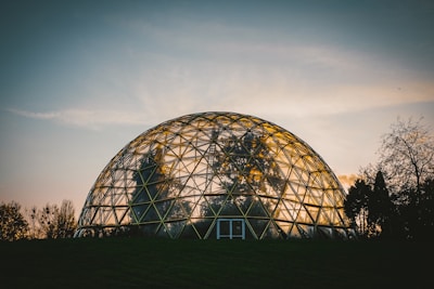 A large geodesic dome made of a metal framework and clear panels is situated in a natural setting. The structure houses plants and trees, partially visible through the transparent surface. The sky is in the background, with soft lighting that suggests either sunrise or sunset.