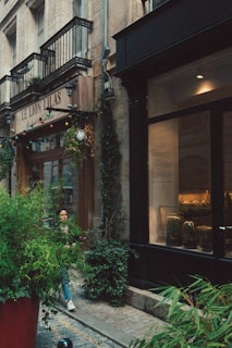 A quaint street scene featuring the exterior of a rustic building with a sign reading 'Le Lion Lilas.' The facade is adorned with small balconies and decorative lighting. Lush green plants in large pots flank both sides of the entrance. The ambiance suggests a cozy, inviting atmosphere. A person is walking on the cobblestone street beside the building, partially obscured by the greenery.