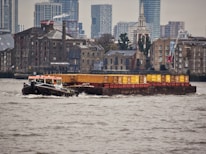 A tugboat is towing a barge loaded with shipping containers across a river. In the background, there is a mix of old brick buildings and modern high-rise structures. The weather appears to be overcast, and the water is slightly choppy.