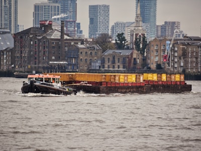 A tugboat is towing a barge loaded with shipping containers across a river. In the background, there is a mix of old brick buildings and modern high-rise structures. The weather appears to be overcast, and the water is slightly choppy.
