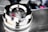 A shiny chrome car part features a cap with an embossed horse emblem and vertical red, white, and blue stripes. The polished surface reflects its surroundings, indicating cleanliness and attention to detail. The background is slightly blurred, focusing attention on the central object.