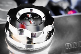 A shiny chrome car part features a cap with an embossed horse emblem and vertical red, white, and blue stripes. The polished surface reflects its surroundings, indicating cleanliness and attention to detail. The background is slightly blurred, focusing attention on the central object.