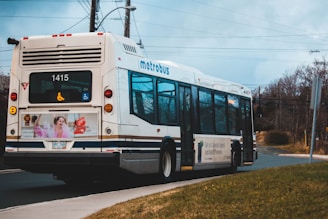 A metro bus with the number 1415 is traveling on a road. The bus displays symbols indicating wheelchair accessibility. The advertisement on the back features a person and colorful graphics. Trees, power lines, and a cloudy sky are in the background.