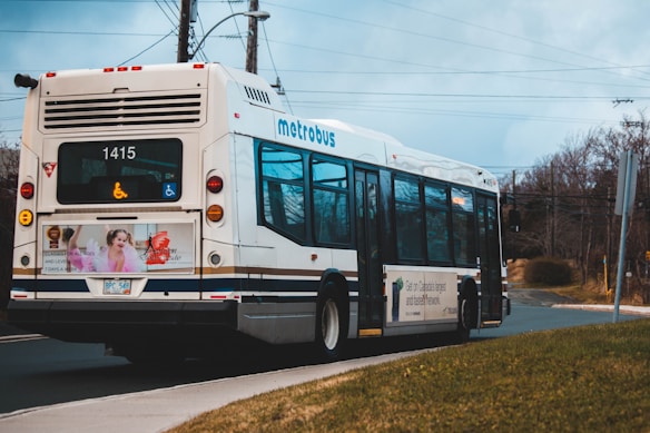 A metro bus with the number 1415 is traveling on a road. The bus displays symbols indicating wheelchair accessibility. The advertisement on the back features a person and colorful graphics. Trees, power lines, and a cloudy sky are in the background.