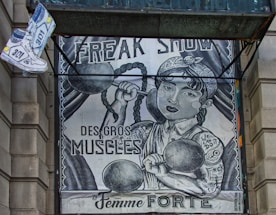 The image depicts an artistic poster resembling a vintage circus or freak show advertisement. It features a strong woman with braided hair lifting dumbbells, with phrases like 'FREAK SHOW,' 'DES GROS MUSCLES,' and 'La Femme FORTE' prominently displayed. There's also a floating pair of sneakers on the top left.