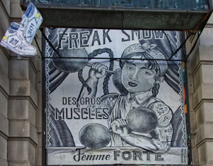 The image depicts an artistic poster resembling a vintage circus or freak show advertisement. It features a strong woman with braided hair lifting dumbbells, with phrases like 'FREAK SHOW,' 'DES GROS MUSCLES,' and 'La Femme FORTE' prominently displayed. There's also a floating pair of sneakers on the top left.