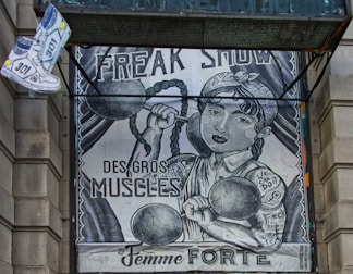 The image depicts an artistic poster resembling a vintage circus or freak show advertisement. It features a strong woman with braided hair lifting dumbbells, with phrases like 'FREAK SHOW,' 'DES GROS MUSCLES,' and 'La Femme FORTE' prominently displayed. There's also a floating pair of sneakers on the top left.