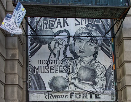 The image depicts an artistic poster resembling a vintage circus or freak show advertisement. It features a strong woman with braided hair lifting dumbbells, with phrases like 'FREAK SHOW,' 'DES GROS MUSCLES,' and 'La Femme FORTE' prominently displayed. There's also a floating pair of sneakers on the top left.