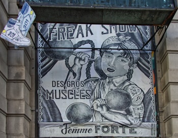 The image depicts an artistic poster resembling a vintage circus or freak show advertisement. It features a strong woman with braided hair lifting dumbbells, with phrases like 'FREAK SHOW,' 'DES GROS MUSCLES,' and 'La Femme FORTE' prominently displayed. There's also a floating pair of sneakers on the top left.