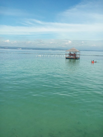 A serene seascape with calm turquoise waters, a small wooden gazebo with a thatched roof on stilts, and a person in a life jacket swimming nearby. The sky is mostly clear with a few clouds, and the horizon shows a distant landmass.