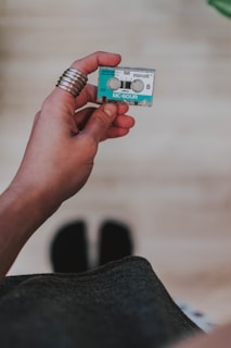 A hand is holding a small, retro cassette tape labeled with 'maxell' and 'MC-60UR'. The hand has several metallic rings on the fingers, and the background is out of focus, possibly showing a blurred floor.