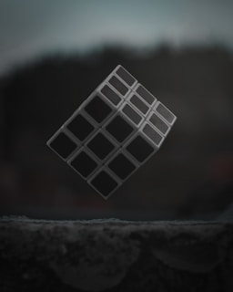A three-dimensional cube with a grid-like pattern levitates against a dark, blurred background. The surface below is textured and appears to be in low light.