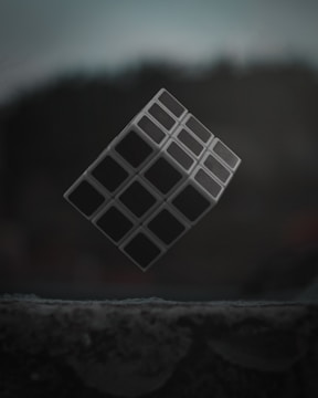 A three-dimensional cube with a grid-like pattern levitates against a dark, blurred background. The surface below is textured and appears to be in low light.