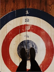 A pair of feet dressed in black shoes stand on a large, wooden target painted with concentric circles. The target features numbered sections in colors of blue, red, and white, and is set on a wood-grain floor.