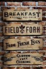 Four wooden signs with black and red text are mounted on a brick wall. The signs read: 'Breakfast Served All Day', 'Field to Fork', 'Farm Fresh Eggs', and 'Gluten Free Cooking', each with distinct fonts and designs. There are also illustrated hands pointing on the last sign.
