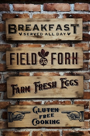 Four wooden signs with black and red text are mounted on a brick wall. The signs read: 'Breakfast Served All Day', 'Field to Fork', 'Farm Fresh Eggs', and 'Gluten Free Cooking', each with distinct fonts and designs. There are also illustrated hands pointing on the last sign.