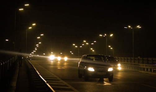 An empty road under night lighting with a row of street lamps illuminating the scene. A few cars with headlights on are visible, contributing to the dim lighting. The road has clear lane markings and is bordered by safety railings.