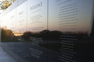 A polished black stone wall displays engraved text featuring principles such as Integrity, Service, and Excellence. The reflective surface captures a scenic view of trees and the horizon in the background, suggesting an outdoor memorial or monument.