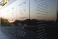A polished black stone wall displays engraved text featuring principles such as Integrity, Service, and Excellence. The reflective surface captures a scenic view of trees and the horizon in the background, suggesting an outdoor memorial or monument.