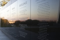 A polished black stone wall displays engraved text featuring principles such as Integrity, Service, and Excellence. The reflective surface captures a scenic view of trees and the horizon in the background, suggesting an outdoor memorial or monument.