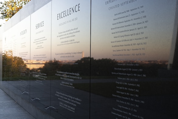 A polished black stone wall displays engraved text featuring principles such as Integrity, Service, and Excellence. The reflective surface captures a scenic view of trees and the horizon in the background, suggesting an outdoor memorial or monument.