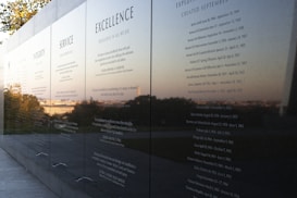 A polished black stone wall displays engraved text featuring principles such as Integrity, Service, and Excellence. The reflective surface captures a scenic view of trees and the horizon in the background, suggesting an outdoor memorial or monument.