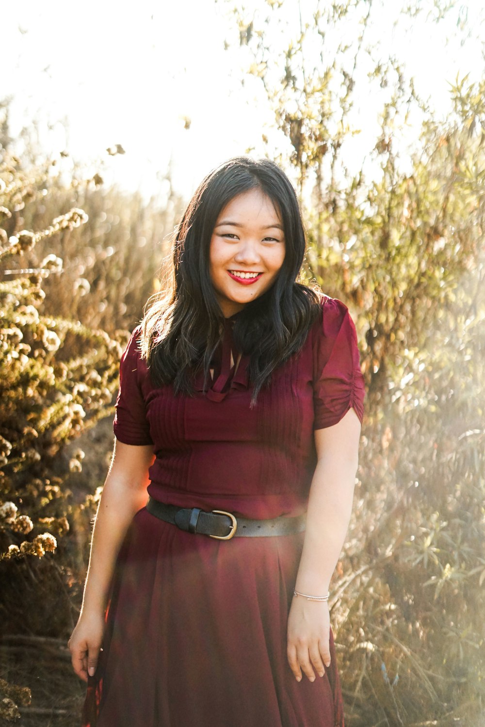 smiling woman standing in trees wearing maroon short-sleeved dress during daytime