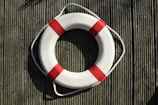 A white lifebuoy with red bands is placed on a textured wooden surface, casting a shadow in the sunlight. The lifebuoy is attached to a rope and appears as if mounted for visibility or use in an emergency.