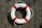 A white lifebuoy with red bands is placed on a textured wooden surface, casting a shadow in the sunlight. The lifebuoy is attached to a rope and appears as if mounted for visibility or use in an emergency.