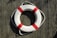 A white lifebuoy with red bands is placed on a textured wooden surface, casting a shadow in the sunlight. The lifebuoy is attached to a rope and appears as if mounted for visibility or use in an emergency.