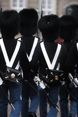 A group of uniformed guards marching, wearing tall black fur hats and formal military attire with white cross-belts and blue trousers. They carry rifles and have decorative pouches on their backs.