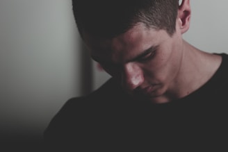 A moody, cinematic shot of a lone young man standing in shadow, gazing sideways with a thoughtful, melancholic expression.