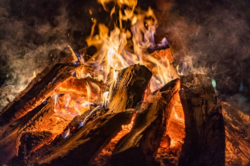 A large arrangement of wooden logs is burning with bright, lively flames. The yellow, orange, and blue hues of the flames create an intense contrast against the dark, smoky background. The logs appear charred and are surrounded by a halo of heat and light.