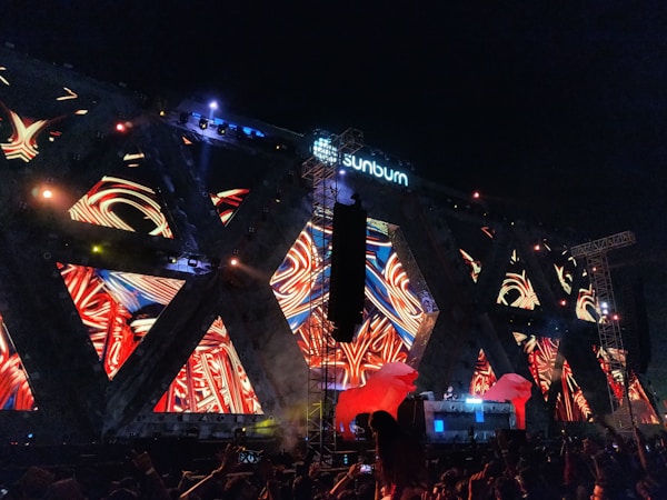 Bright, colorful lights and vibrant patterns are displayed on a large stage structure at a nighttime outdoor music festival. The structure features a mix of abstract, swirling designs with a central DJ booth, surrounded by enthusiastic crowd members. The brand 'sunburn' is visible at the top.
