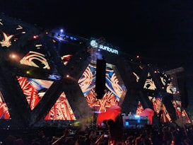 Bright, colorful lights and vibrant patterns are displayed on a large stage structure at a nighttime outdoor music festival. The structure features a mix of abstract, swirling designs with a central DJ booth, surrounded by enthusiastic crowd members. The brand 'sunburn' is visible at the top.