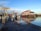 A dock area with several boats moored, situated in front of a large building with a red roof. The scene features calm water reflecting the sky and surroundings, with piers and walkways visible. The sign notes the location as Steveston Gulf, managed by the Steveston Harbour Authority.