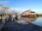 A dock area with several boats moored, situated in front of a large building with a red roof. The scene features calm water reflecting the sky and surroundings, with piers and walkways visible. The sign notes the location as Steveston Gulf, managed by the Steveston Harbour Authority.