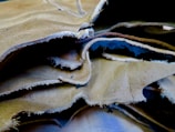 A close-up view of several overlapping sheets of beige and light brown leather with frayed edges. The texture of the leather is visible, with slight wrinkles and natural imperfections.