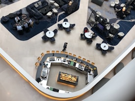 An aerial view of a modern, stylish lounge area featuring a central curved bar with a variety of glasses and bottles. Surrounding this are groups of tables with chairs, where people are engaged in conversation. The space is divided into areas with different styles of seating, including couches and armchairs. The floor is tiled, and the design includes contrasting dark and light areas.