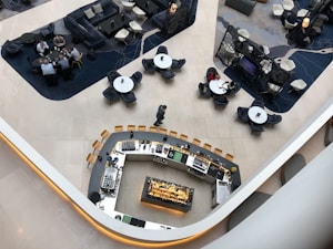 An aerial view of a modern, stylish lounge area featuring a central curved bar with a variety of glasses and bottles. Surrounding this are groups of tables with chairs, where people are engaged in conversation. The space is divided into areas with different styles of seating, including couches and armchairs. The floor is tiled, and the design includes contrasting dark and light areas.